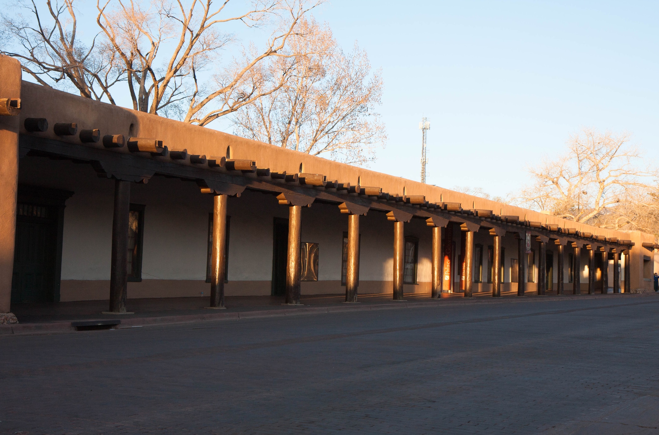 One of the oldest buildings in America, dating back to 1610, the Palace of the Governors hosts a daily Native American Market. Photograph shot at sunset, trees in background, no people visible.